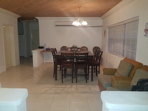 A dining area with a wooden table and chairs, a kitchen in the background, and a window with blinds.