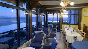 Un restaurant doté de grandes fenêtres donnant sur l’eau, de chaises recouvertes d’un tissu bleu, d’une longue table à manger avec des verres à vin et d’un lustre.
