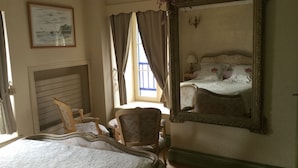 Une chambre à coucher avec un grand miroir décoratif, un lit recouvert d’une literie à motifs floraux, une chaise et une fenêtre avec des rideaux.