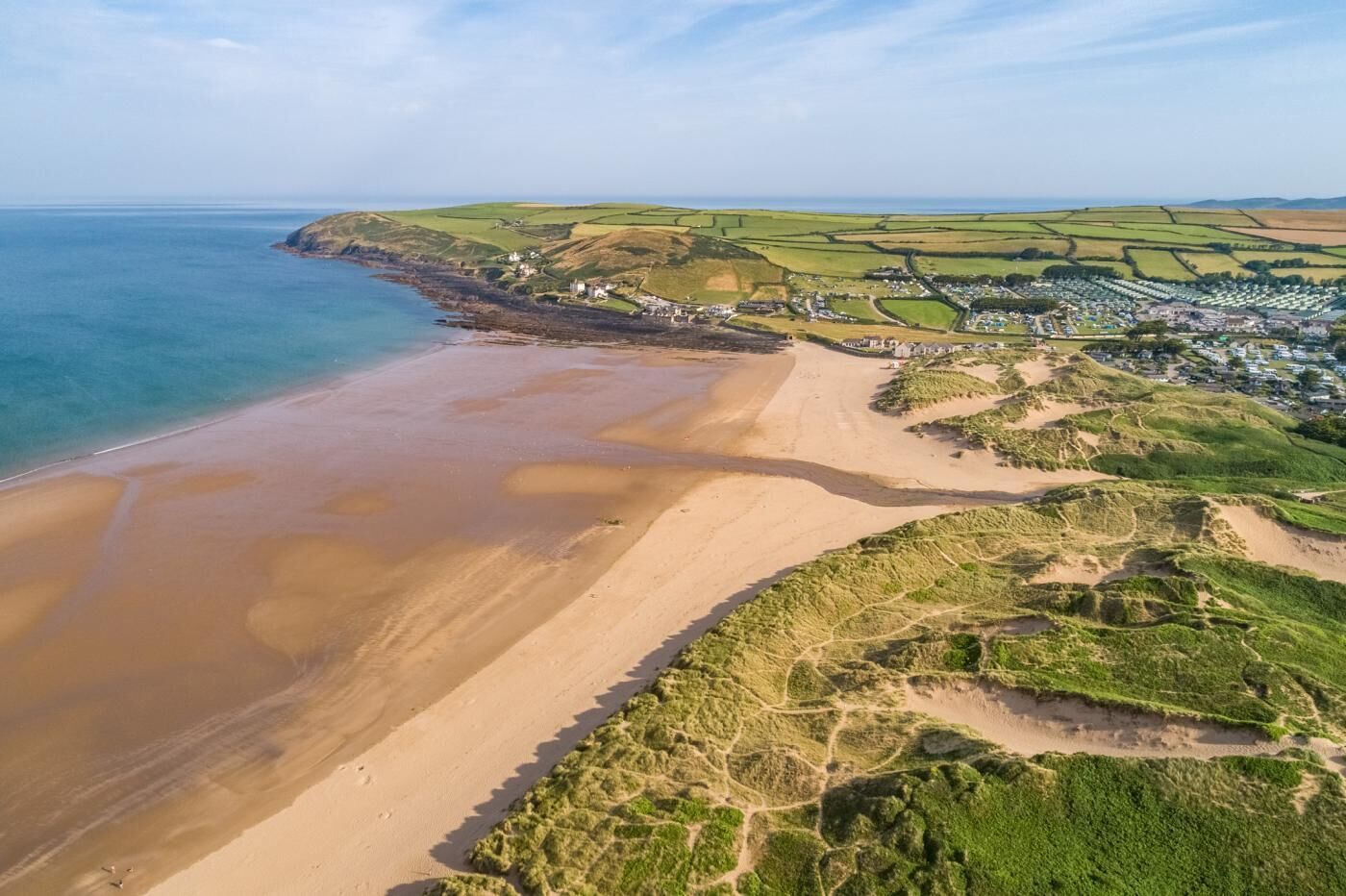 An aerial view of a coastal area with a sandy beach, green fields, and a small settlement with buildings.