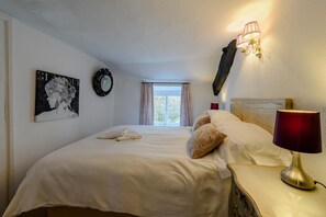 A hotel room with a bed, a bedside lamp, a wall clock, a window with curtains, and a painting on the wall.