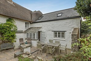 A stone cottage with a thatched roof, a wooden table, and chairs in the courtyard.