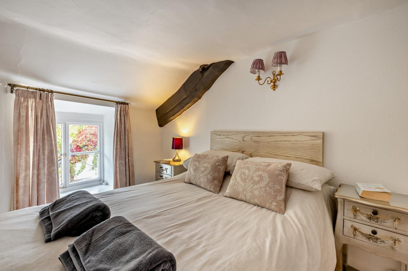Een slaapkamer met een groot bed, een nachtkastje met lamp, een raam met gordijnen en een houten balk aan het plafond.