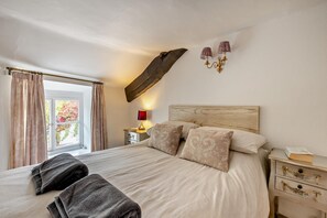A bedroom with a large bed, a nightstand with a lamp, a window with curtains, and a wooden beam on the ceiling.