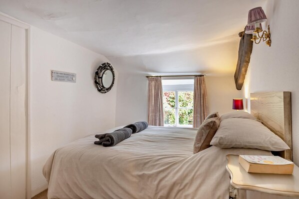 A bedroom with a bed, a bedside table with a book, a wall-mounted lamp, and a window with curtains.