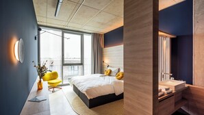 A modern hotel room with a large bed, a bedside table, a chair, a window with curtains, and a bathroom with a sink and mirror. - THE CLOUTH (Cologne)