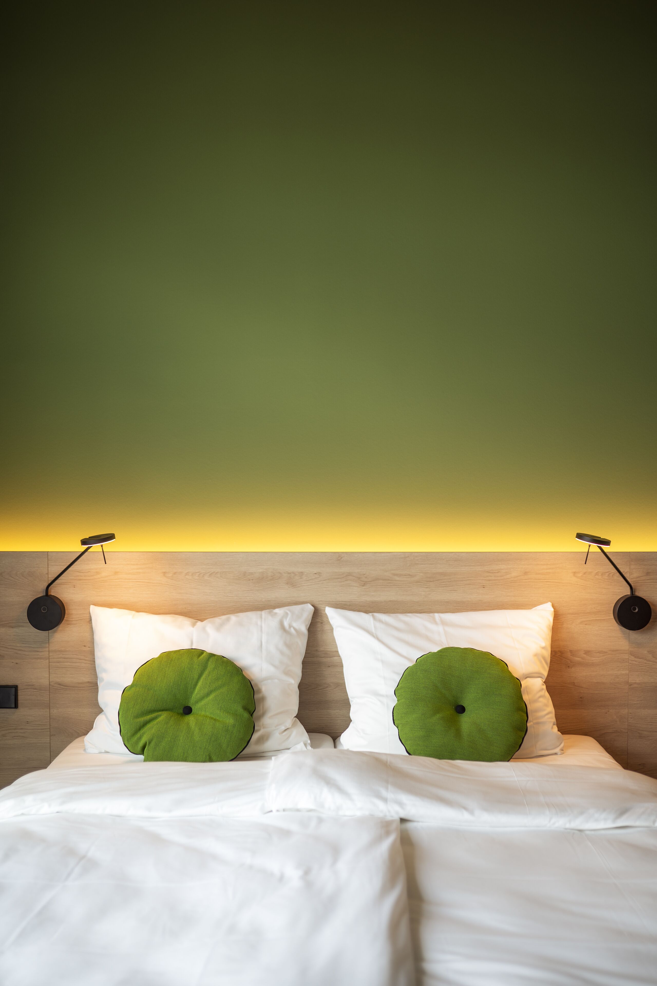 Een bed met witte lakens en twee groene kussens, een houten hoofdbord en twee wandlampen.