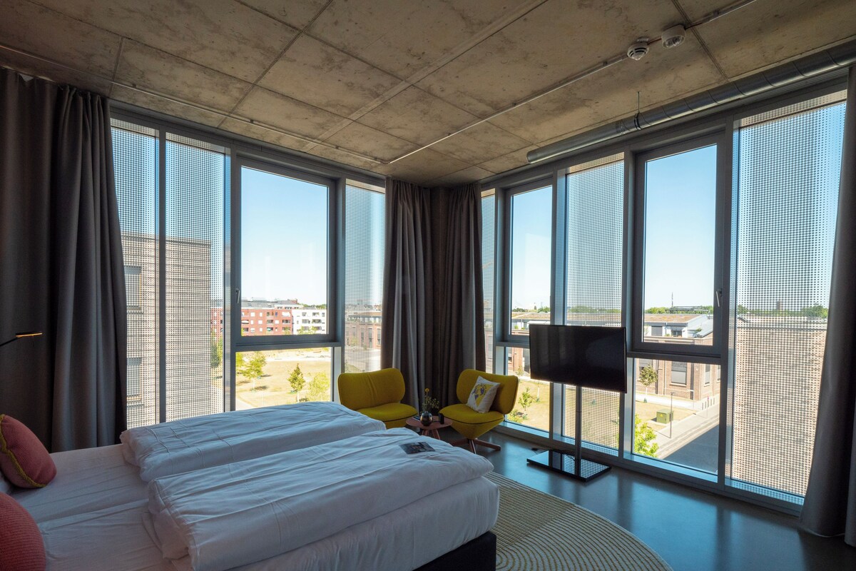 A modern hotel room with a large bed, a yellow armchair, a flat-screen TV, and large windows offering a city view.