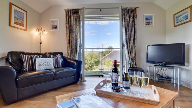 A living room with a leather sofa, a coffee table with wine glasses and a bottle, a TV on a stand, and a balcony with curtains.