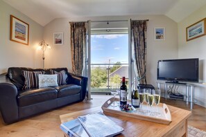 Ein Wohnzimmer mit einer Ledersofa, einem Couchtisch mit Weingläsern und einer Flasche, einem Fernseher auf einem Schrank und einem Balkon mit Vorhängen.
