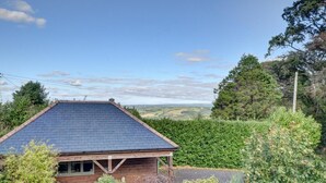 A wooden house with a sloped roof, surrounded by greenery and a view of rolling hills.