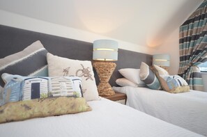A hotel room with two beds, each with white bedding and decorative pillows. There are two wall-mounted lamps with blue shades and a woven side table between the beds.