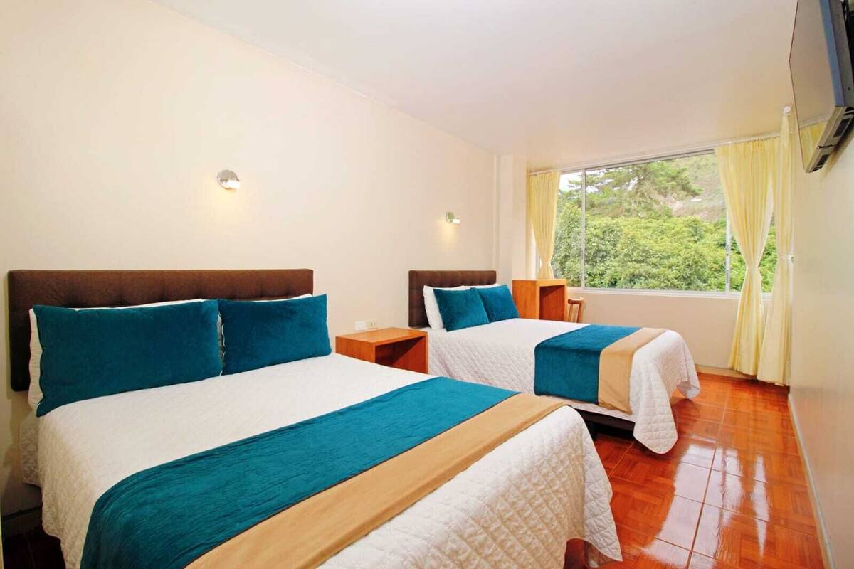 A hotel room with two beds, a window with curtains, and a view of greenery outside.
