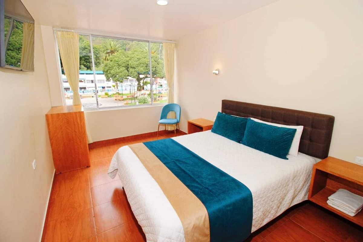 A bedroom with a bed, a chair, a nightstand, a window with curtains, and a view of buildings and trees outside.