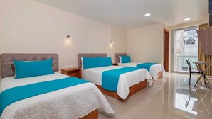 Habitación de hotel con tres camas, cada una con una colcha blanca y azul, cabecero de madera y una mesita de noche pequeña.
