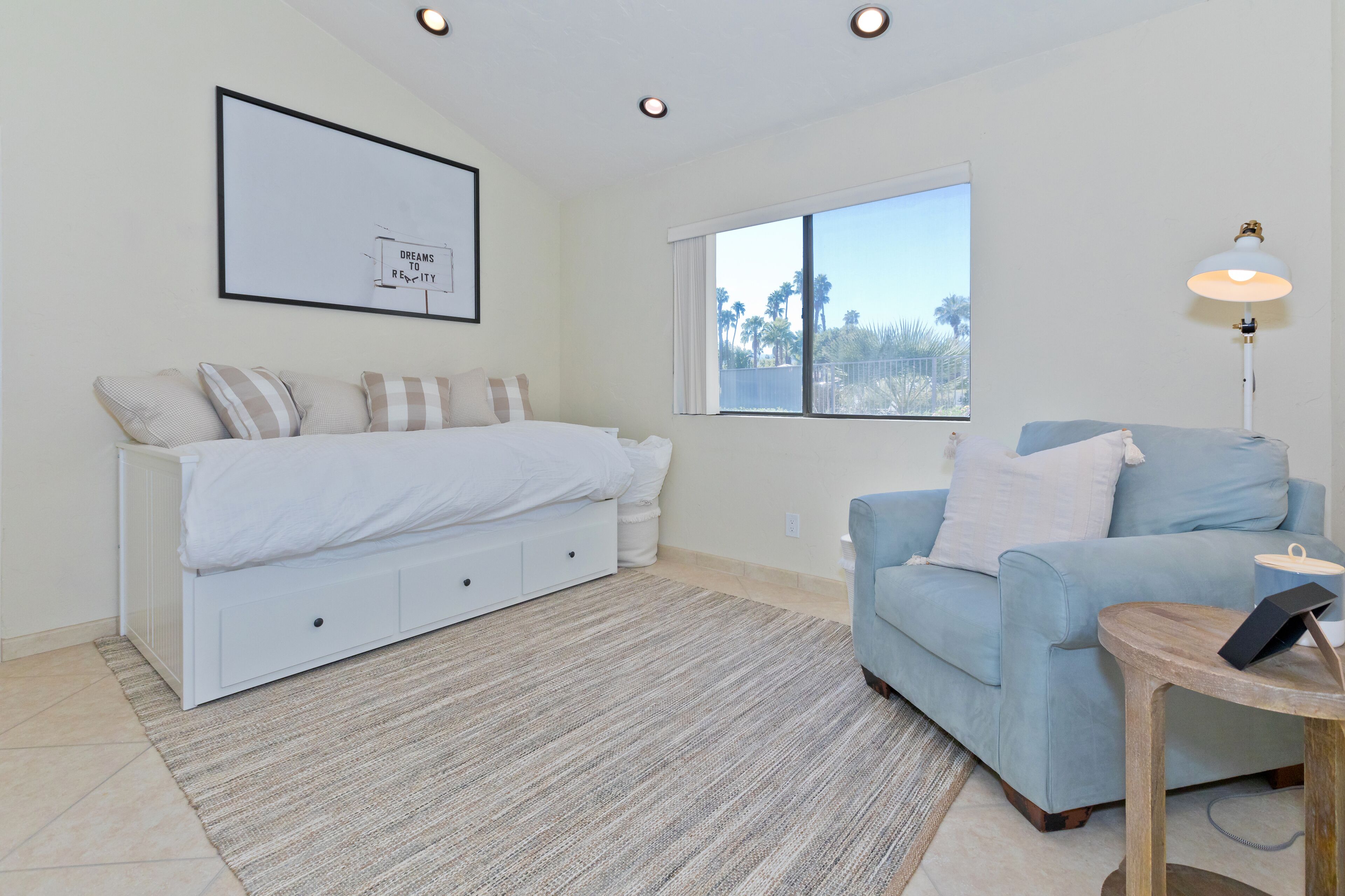 A bedroom with a bed, a blue sofa, a small table, a window with a view of palm trees, and a framed picture on the wall.
