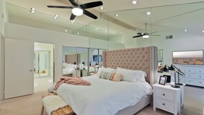 Un dormitorio moderno con una cama grande, una mesita de noche, un ventilador de techo y un espejo amplio.