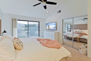 Ein modernes Schlafzimmer mit einem großen Bett, einem Deckenventilator, einer Glastür, die auf einen Balkon führt, einer weißen Kommode und einem großen Spiegel, der den Raum widerspiegelt.