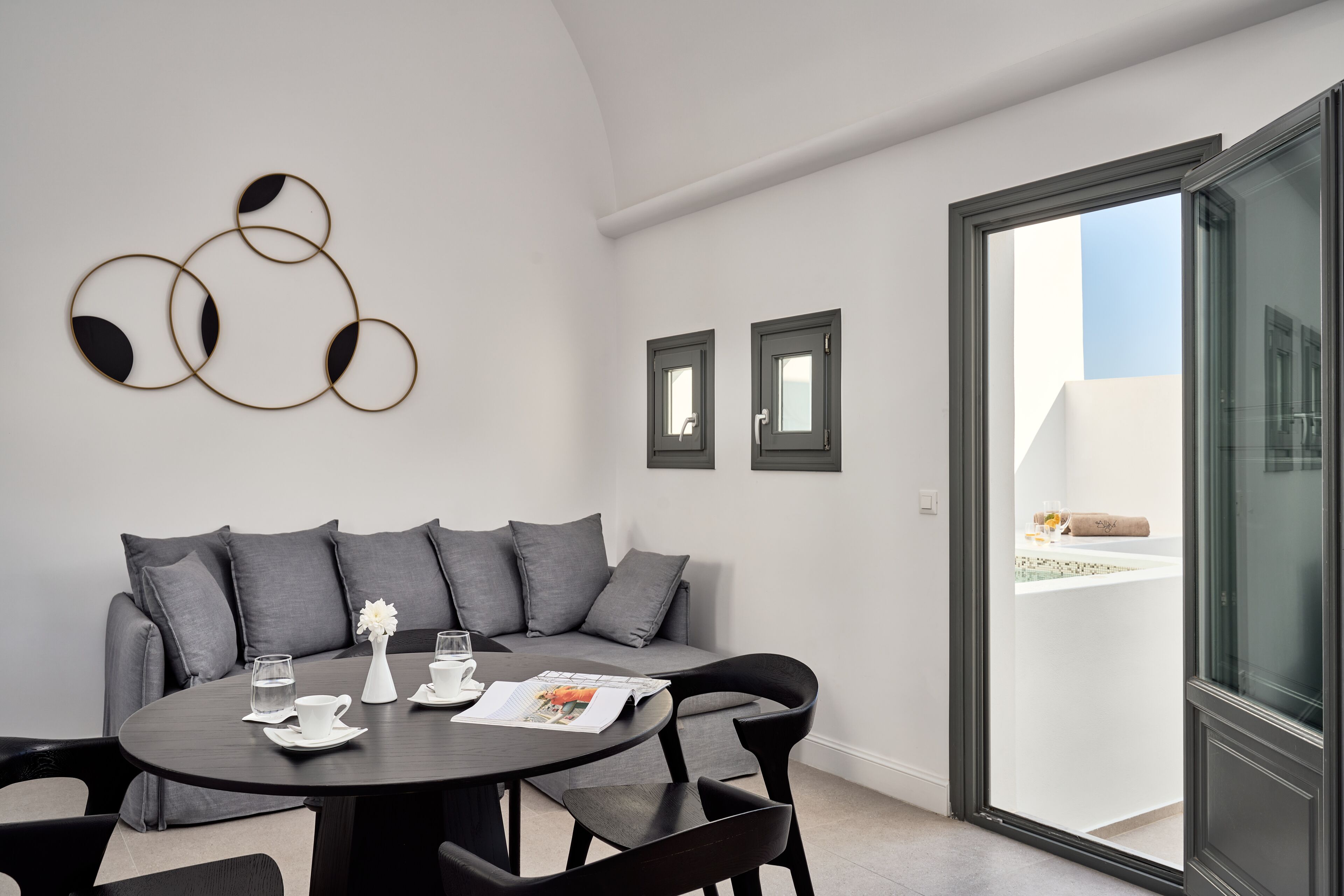 Un salón moderno con una mesa redonda, un sofá gris y un ventanal de vidrio que da a un espacio exterior.
