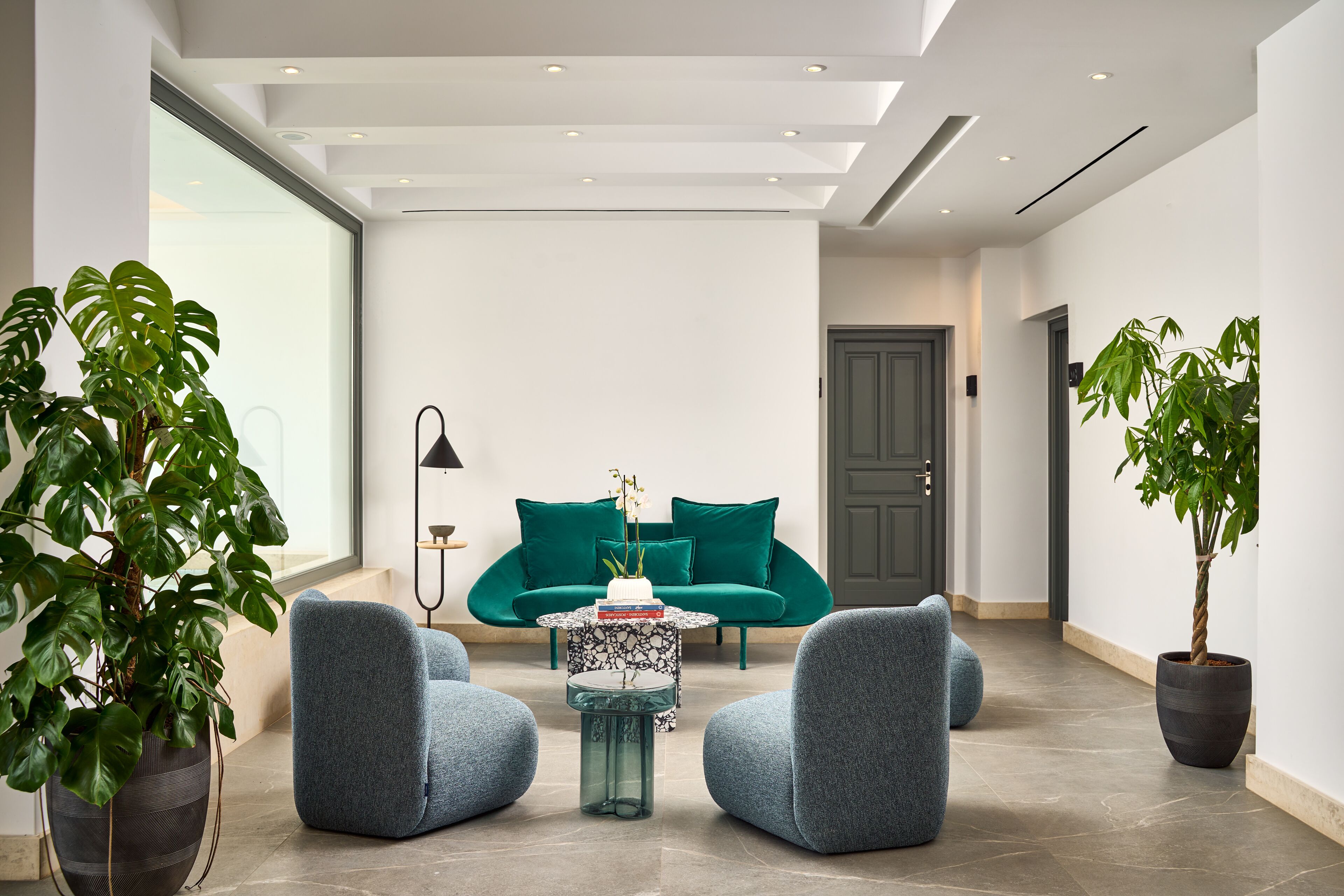 A modern living room with a teal sofa, two armchairs, a glass coffee table, and potted plants.