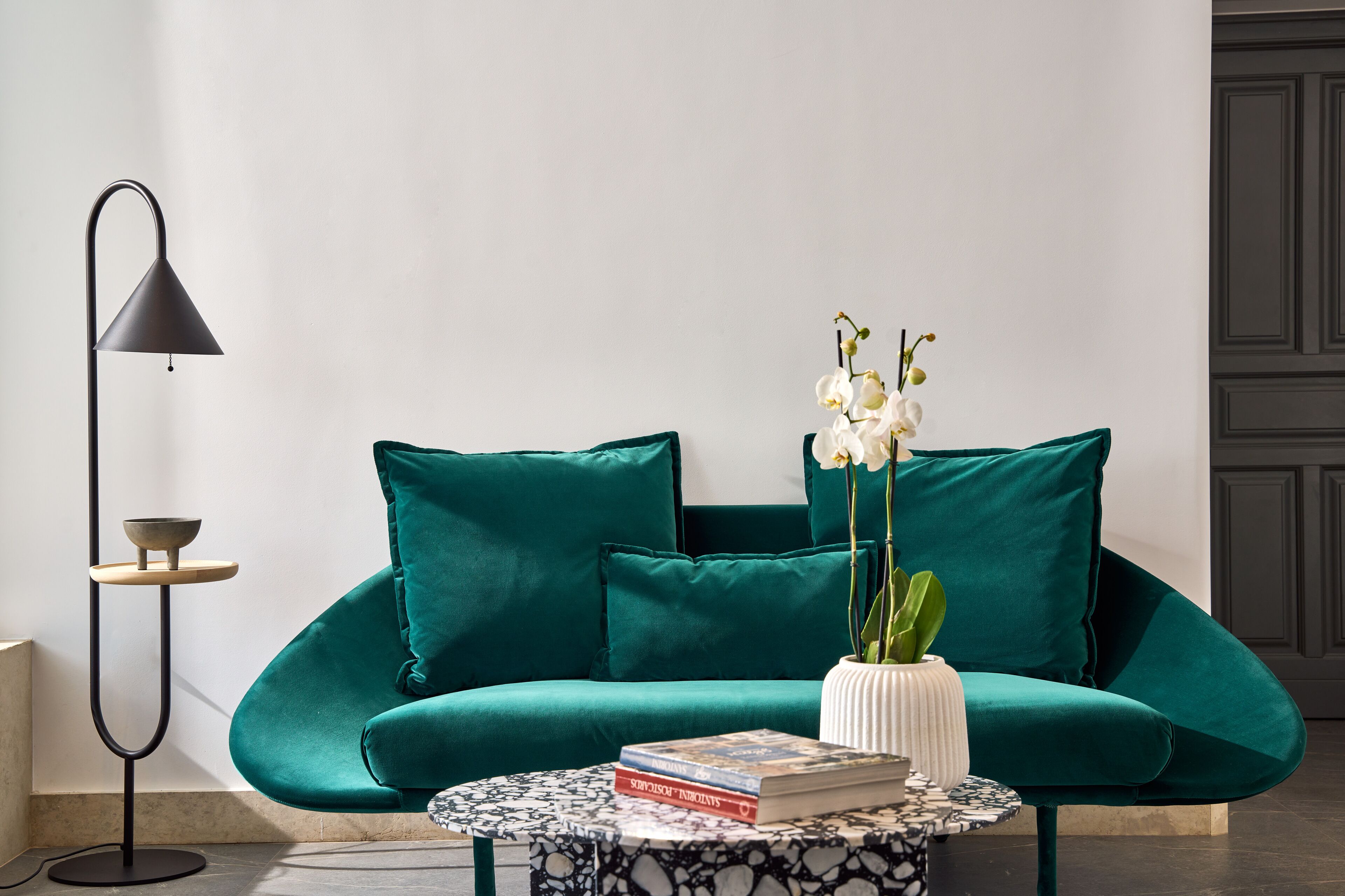A teal sofa with matching cushions, a small round coffee table with books and a vase of white orchids, and a black floor lamp.