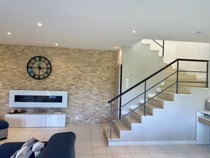 A modern living room with a brick wall, a large clock, a flat-screen TV, a staircase with a metal railing, and a sofa.