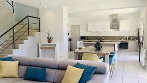 A modern living room with a grey sofa, a dining area, and a kitchen with white cabinets and a stainless steel range hood. - La Gaude, villa 6 people-garden-pool-open view in a quiet (La Gaude)