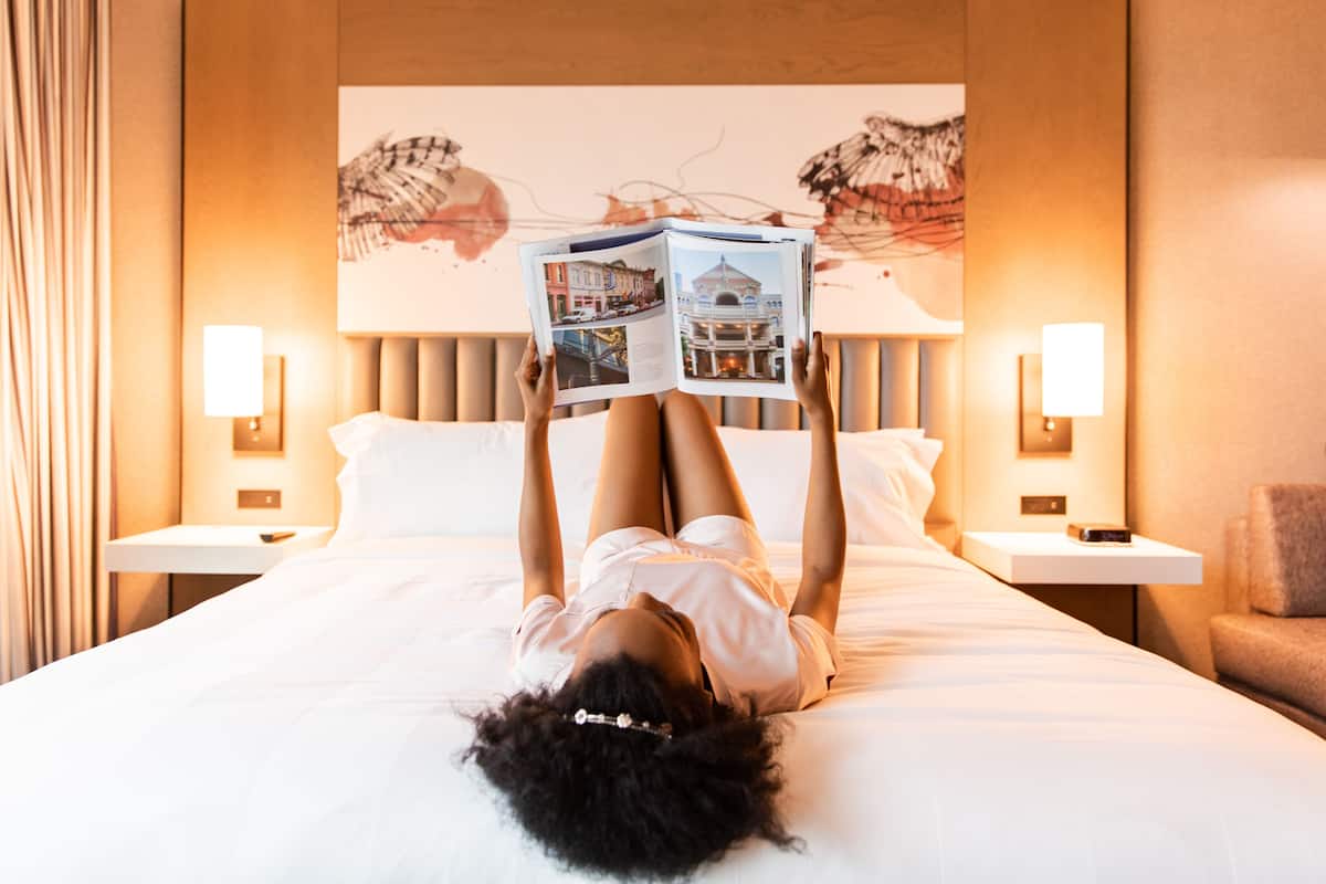 A person lying on a bed in a hotel room, reading a magazine. The room has a bed with white linens, two bedside tables with lamps, and a wall with a large painting.
