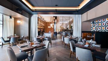 A modern restaurant with multiple dining tables and chairs, a bar area, and a view of the kitchen in the background.