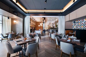 A modern restaurant with multiple dining tables and chairs, a bar area, and a view of the kitchen in the background.