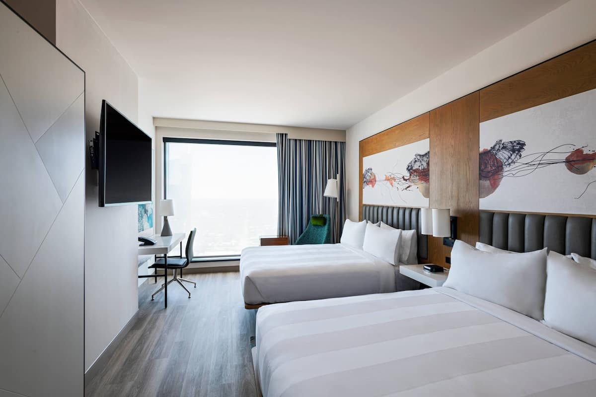 A hotel room with two beds, a desk with a chair, a flat-screen TV, and a large window with curtains.