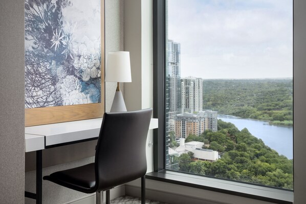 A modern hotel room with a desk, chair, and a large window offering a view of a cityscape and a lake.