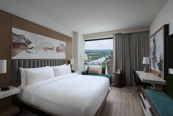 A hotel room with a large bed, a desk, a chair, a view of a cityscape, and a large abstract painting on the wall.