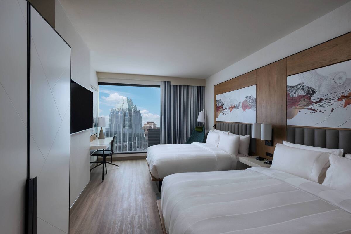 A hotel room with two beds, a desk, and a large window with a cityscape view.