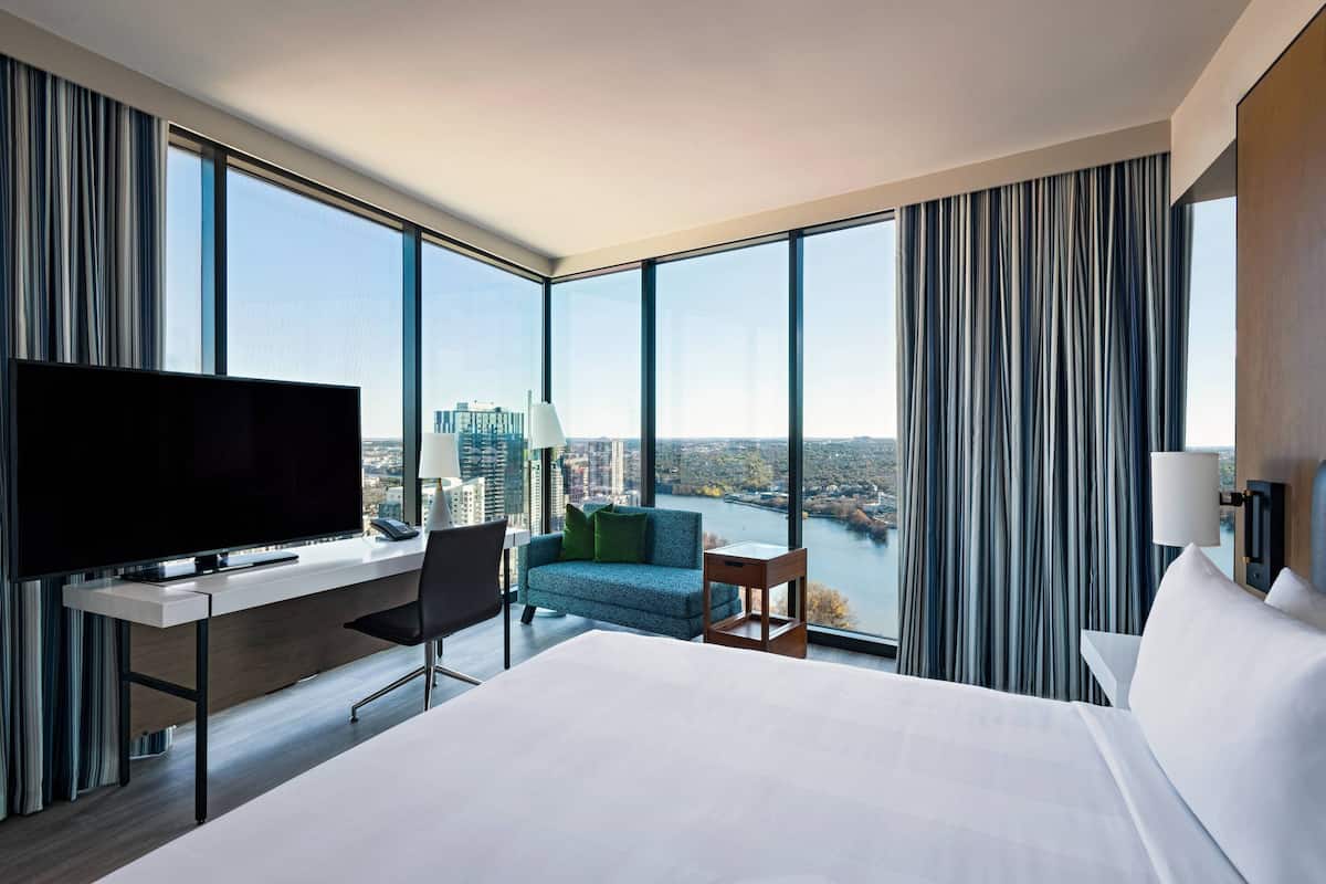 A hotel room with a large bed, a desk with a TV, a chair, a sofa, and a view of a river and cityscape.