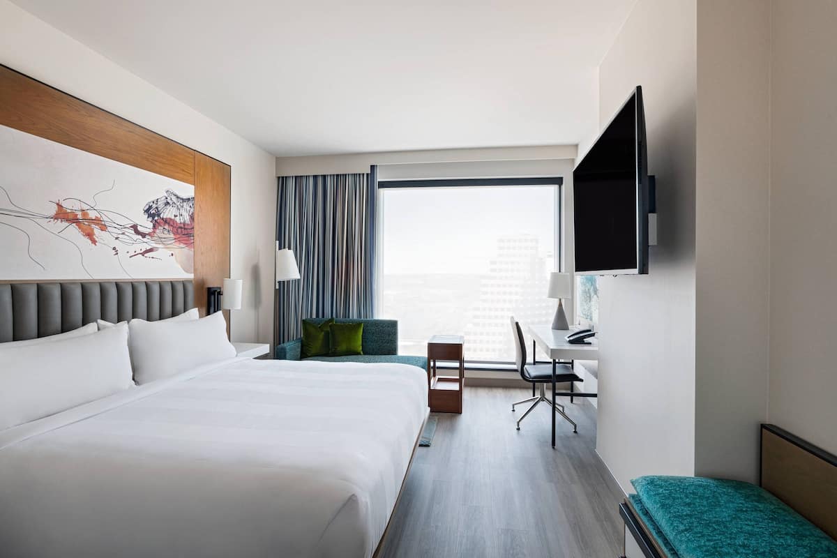 A modern hotel room with a large bed, a desk, a chair, a television, and a window with a city view.