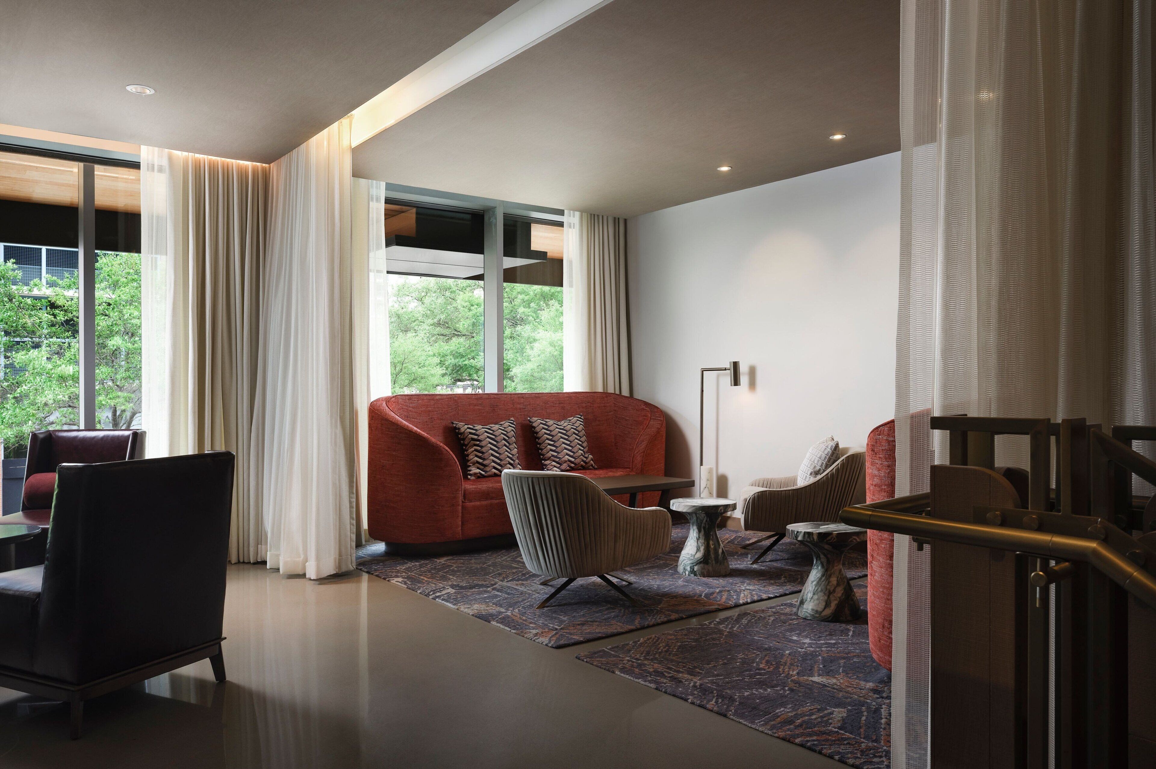 Una sala de estar moderna con un sofá rojo, un sillón gris y una pequeña mesa redonda. Se observa un ventanal con cortinas translúcidas, y una escalera a la derecha.