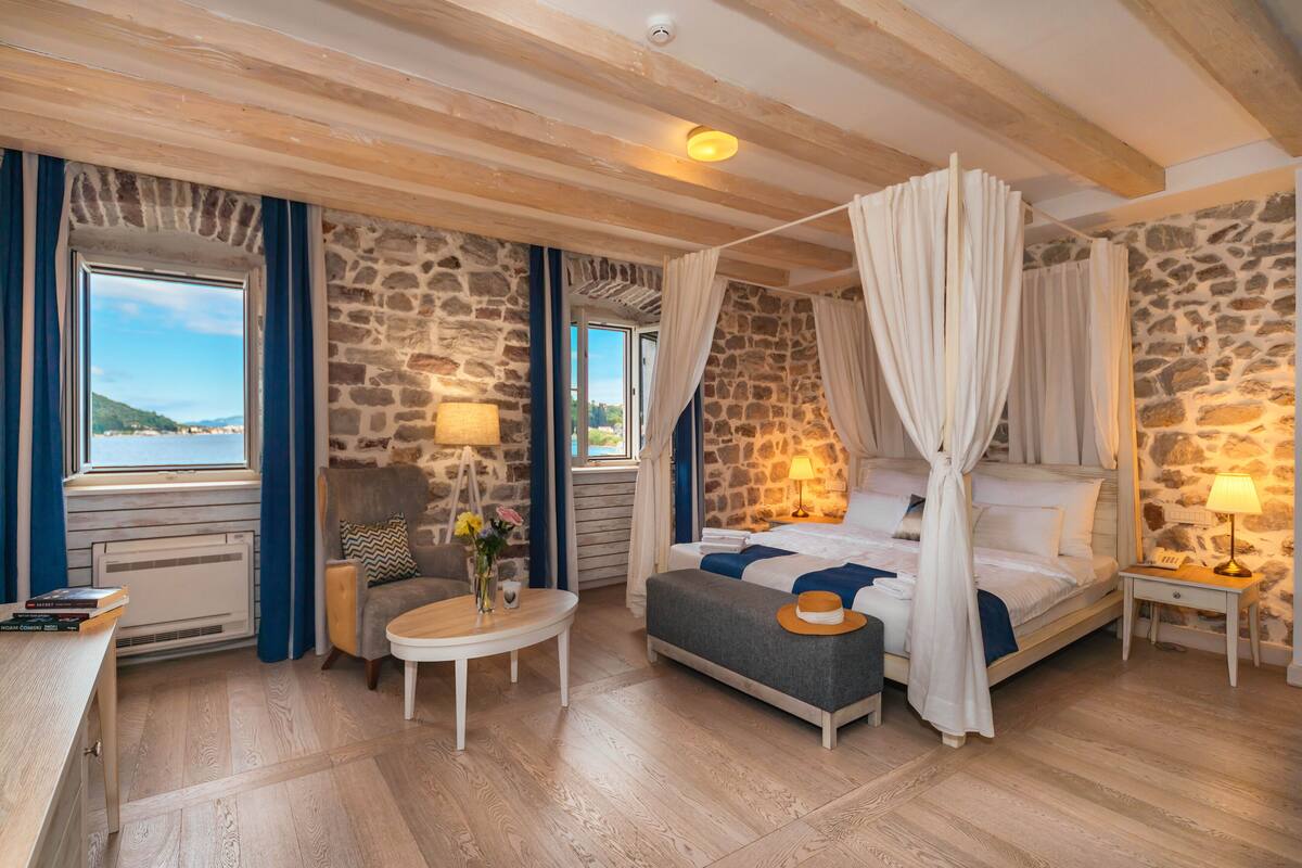 A bedroom with a stone wall, a bed with white curtains, a small table with a lamp, and a view of the sea.