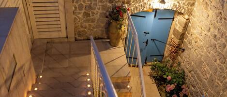 Una escalera de piedra conduce a una puerta azul, rodeada de macetas con plantas y una barandilla.
