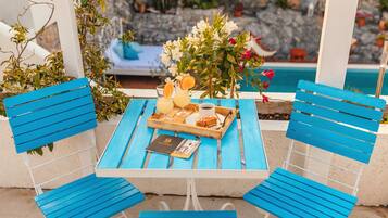 Una mesa de madera azul con una bandeja de comida y bebidas, rodeada de sillas azules, frente a un muro de piedra y una piscina.