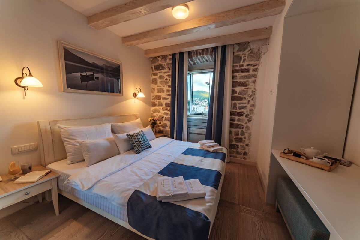 A bedroom with a bed, a nightstand, a desk, a stone wall, and a window with curtains.