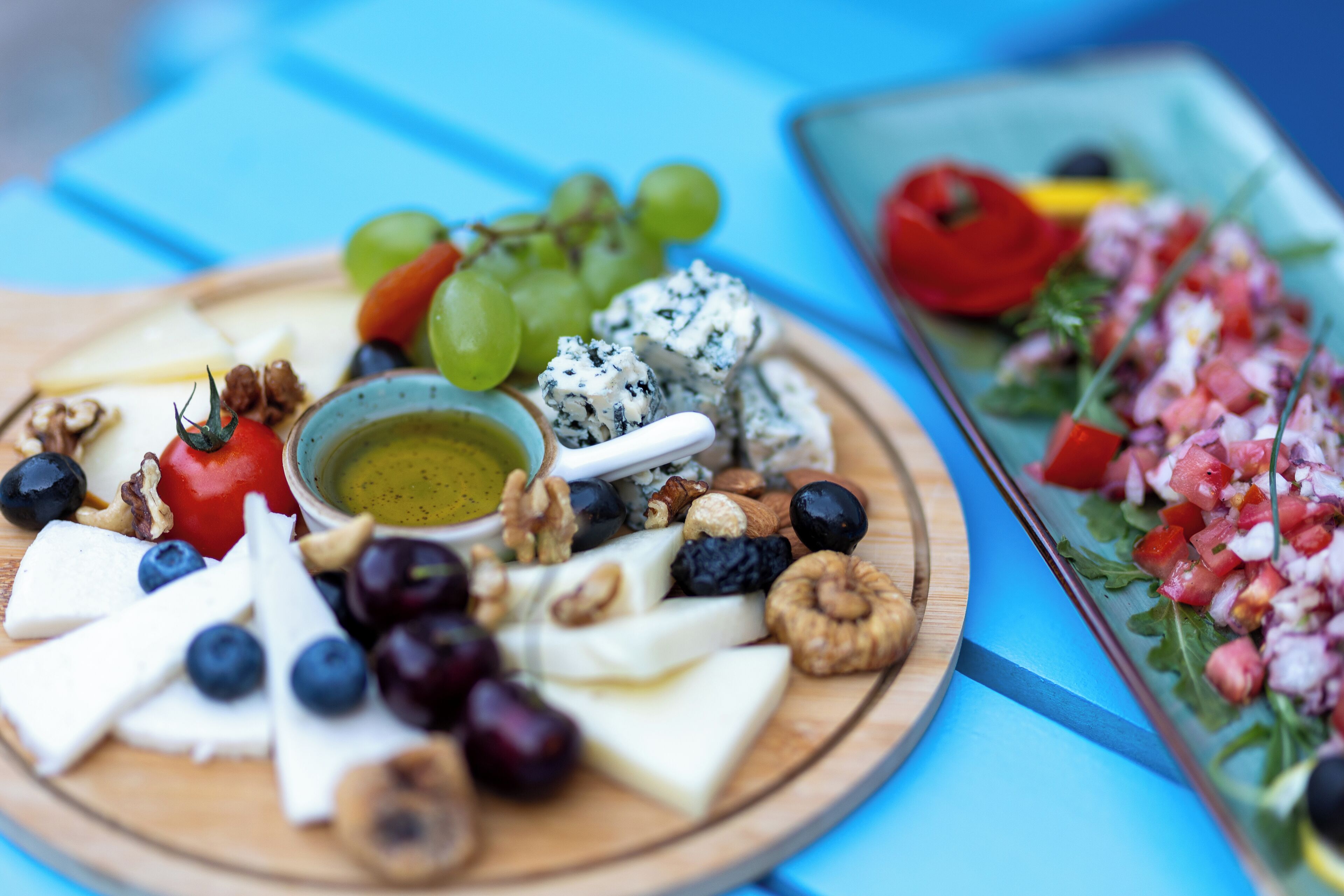 A wooden board with various cheeses, grapes, nuts, and a small bowl of olive oil.