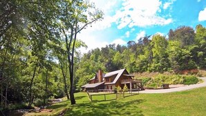 A rustic cabin with a sloped roof, surrounded by a well-maintained lawn and a small stream.