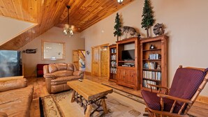 A cozy living room with a wooden ceiling, a leather sofa, a wooden coffee table, and a bookshelf.