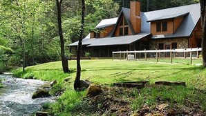 A log cabin with a stone foundation, large windows, and a sloped roof, surrounded by a lush green landscape and a flowing stream.
