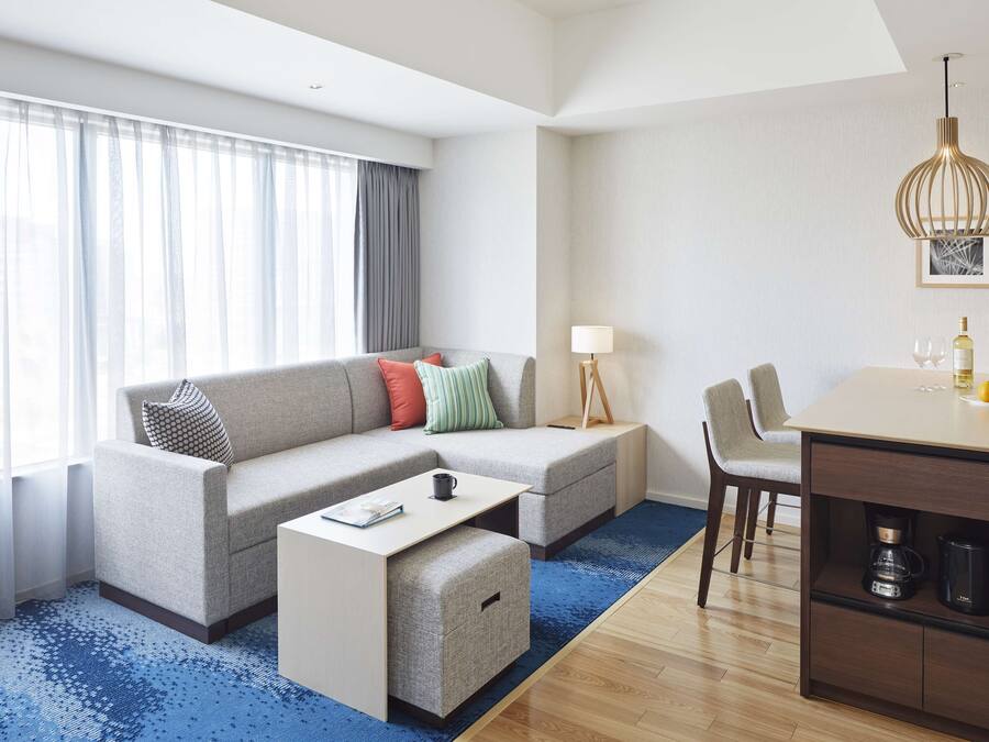 Quarto de hotel moderno com sofá, banquinho, mesa de jantar e cafeteira.