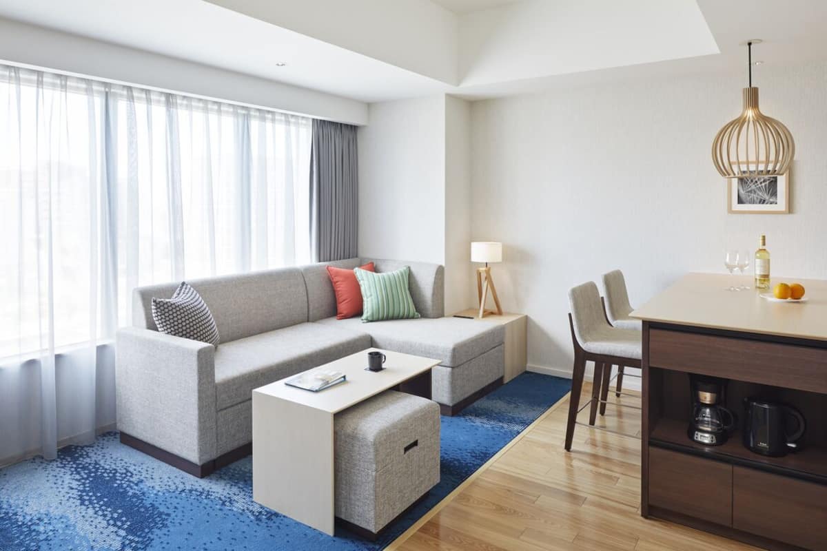A modern hotel room with a sofa, ottoman, dining table, lamp, and coffee maker.