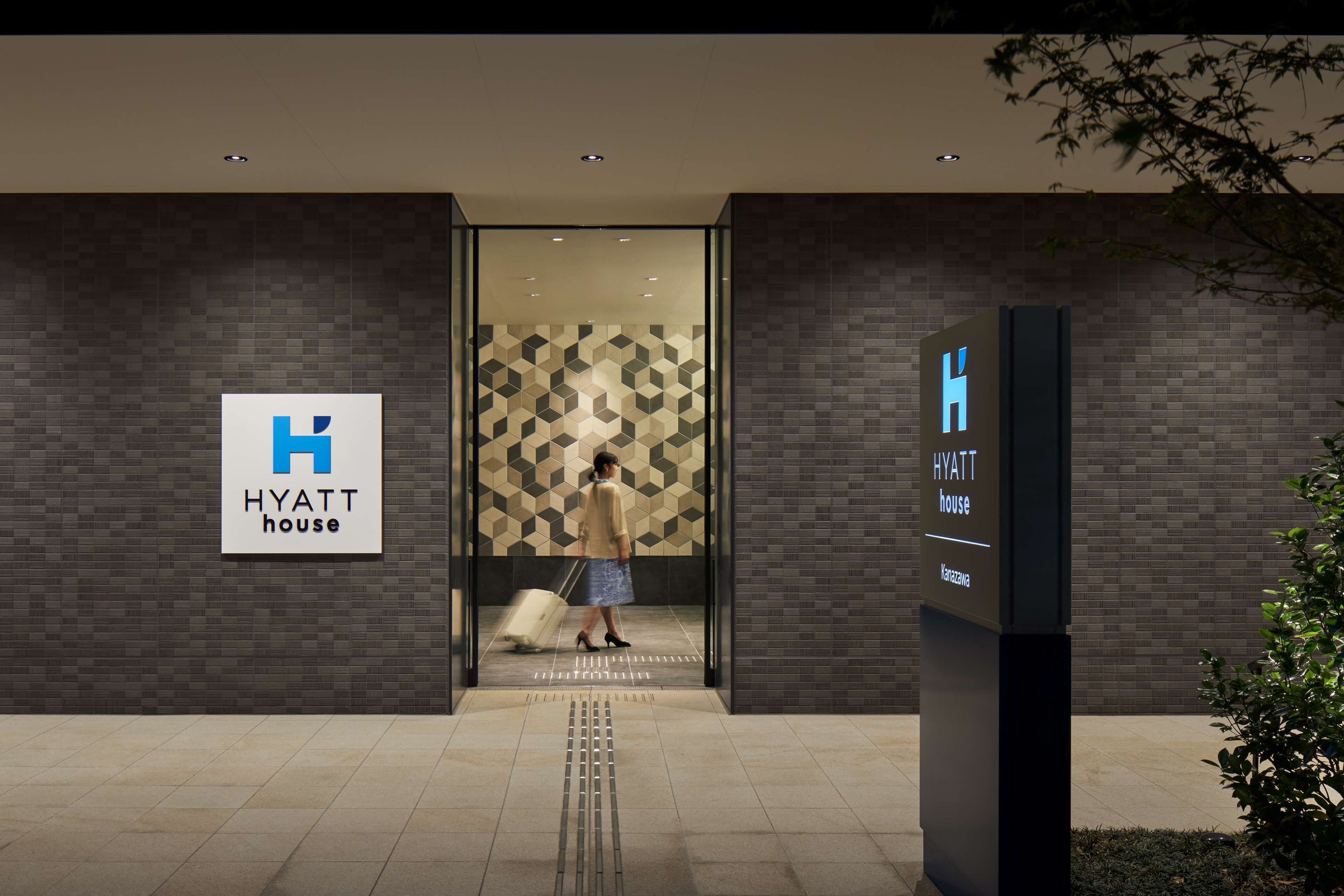 Entrada de um hotel com parede de padrão geométrico, uma placa indicando "HYATT house" e uma pessoa carregando uma mala.