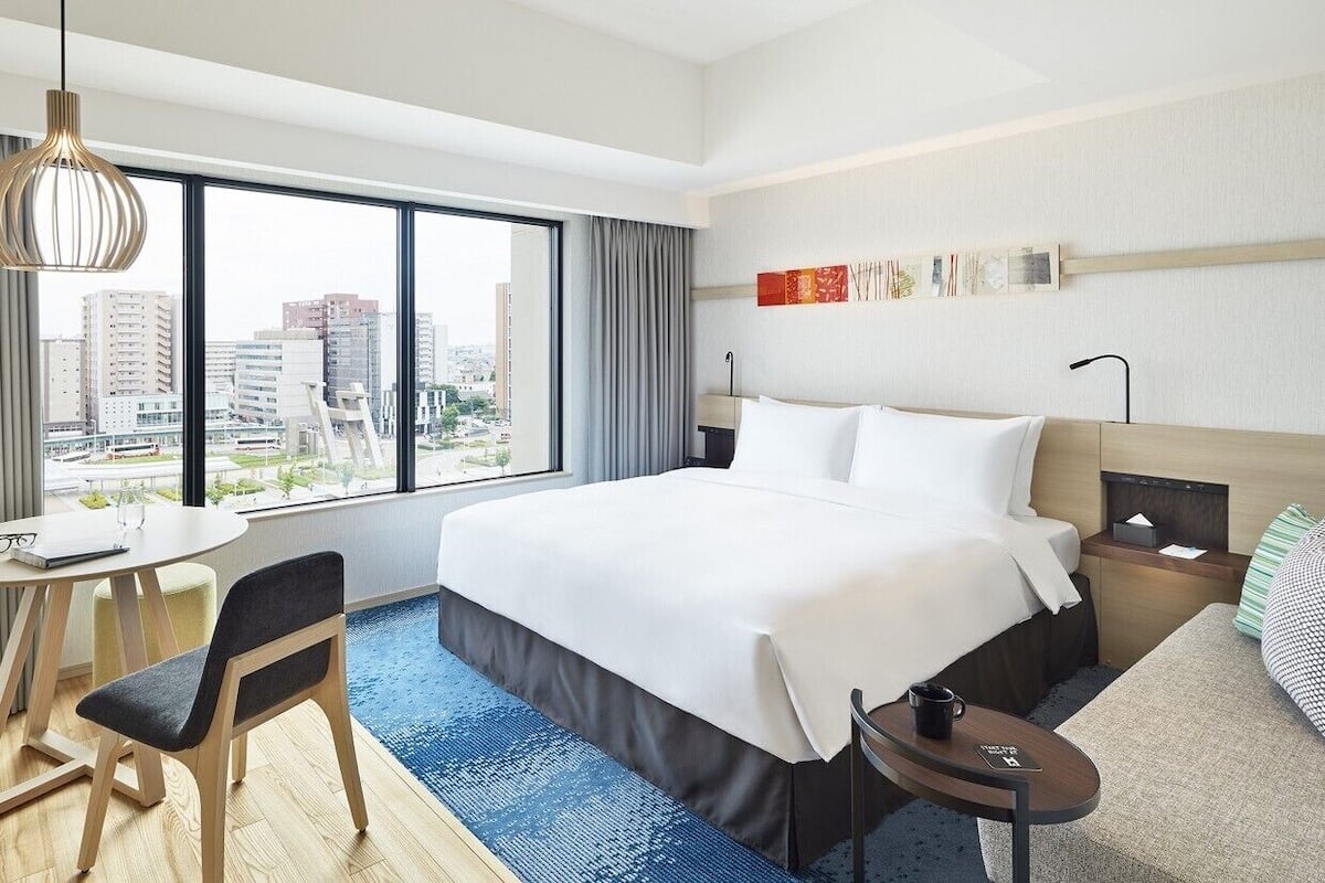 A hotel room with a large bed, a desk with a chair, a small table, and a view of the city.