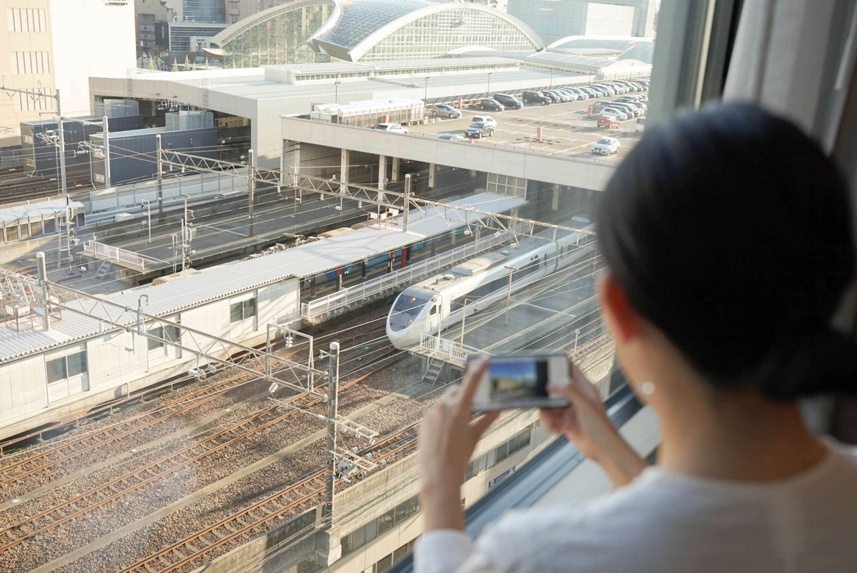 A person taking a photo of a train station with multiple tracks and a modern architectural structure in the background.